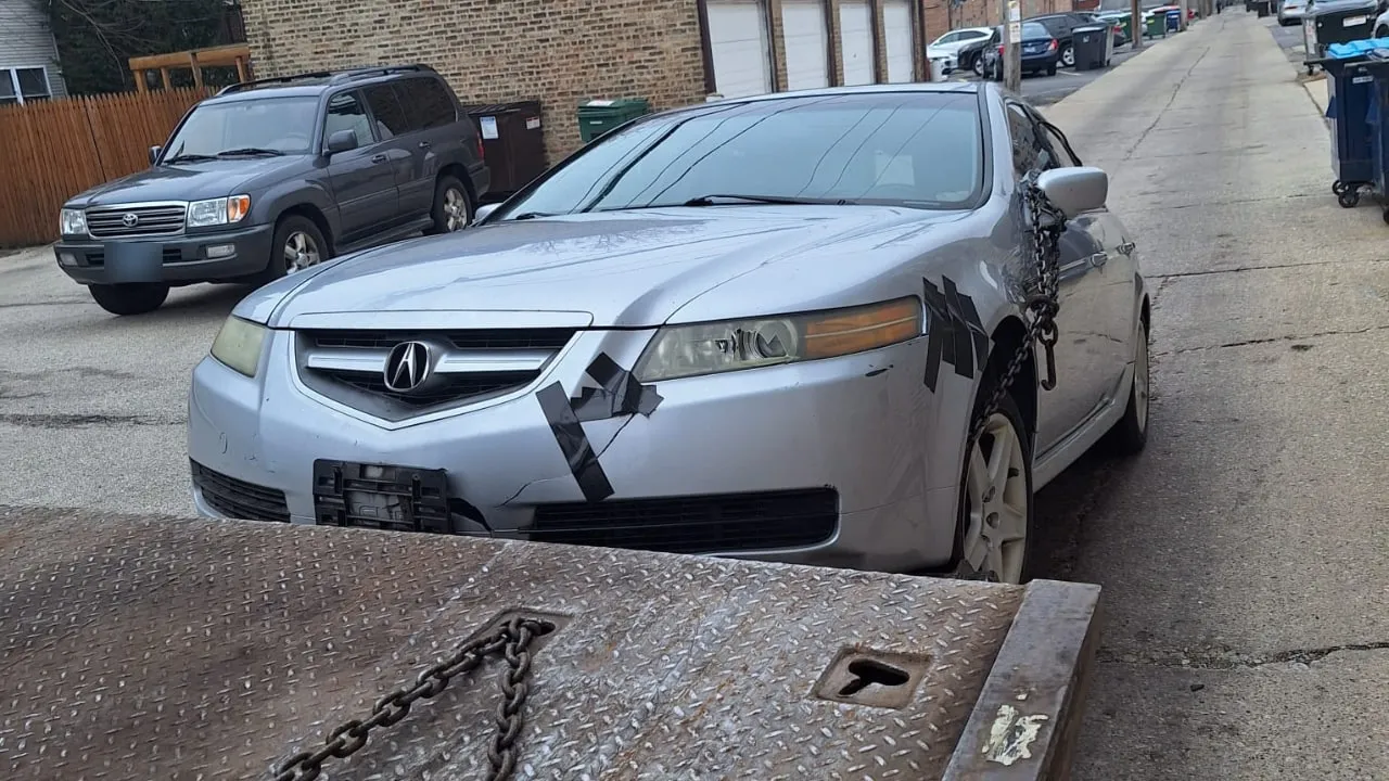 Silver sedan with taped-up front damage, waiting in an alley for the flatbed.